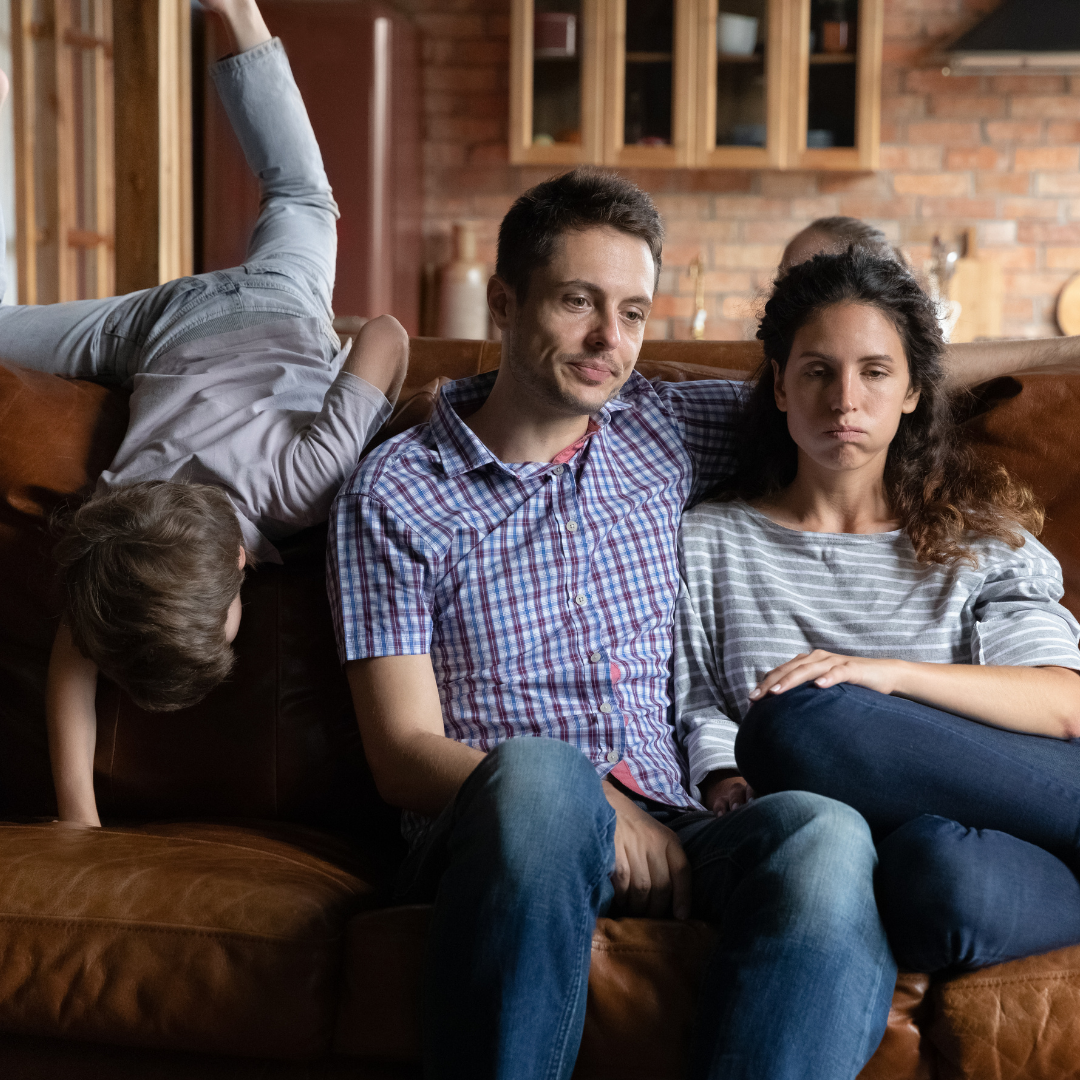 little boy vaulting over couch next to tired dad and exasperated mom