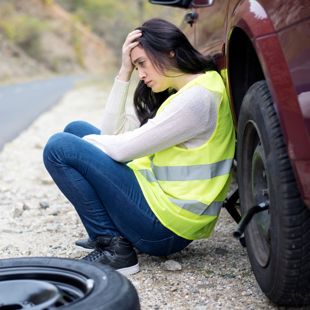 young woman wearing safety vest, sitting with back to car with flat tire